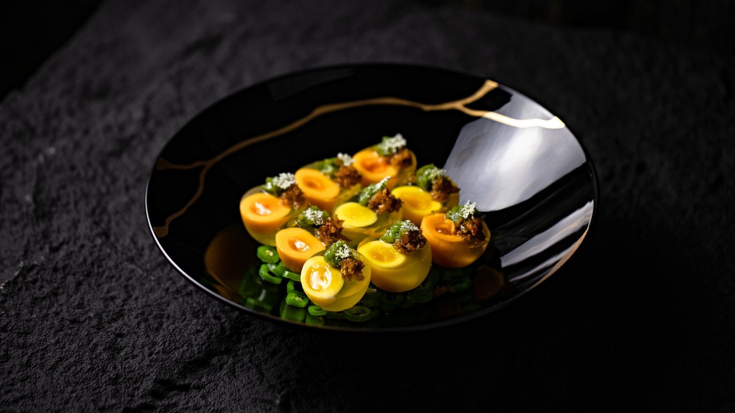Assiette noire veinée d'or, bouchées ambrées dressées à la pince, cuisine à la braise