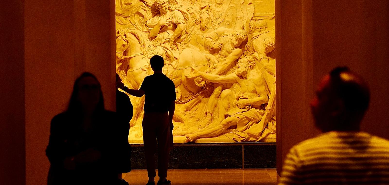 Silhouettes de visiteurs devant un grand relief sculpté baigné de lumière chaude, soir de vernissage